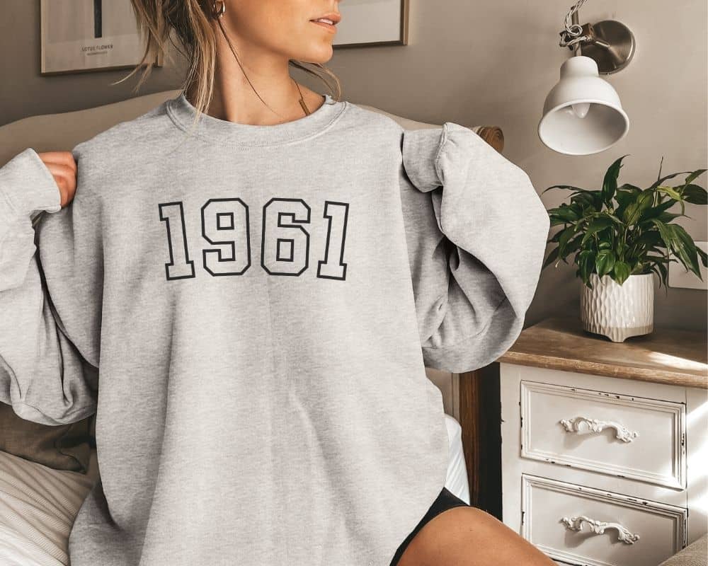 A person wearing a gray sweatshirt with '1961' printed on it, sitting in a room.