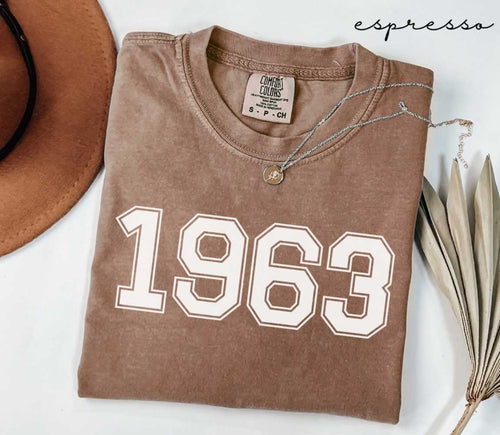 Folded brown (espresso) t-shirt with '1963' print on a white surface, accompanied by a hat and dried leaves.