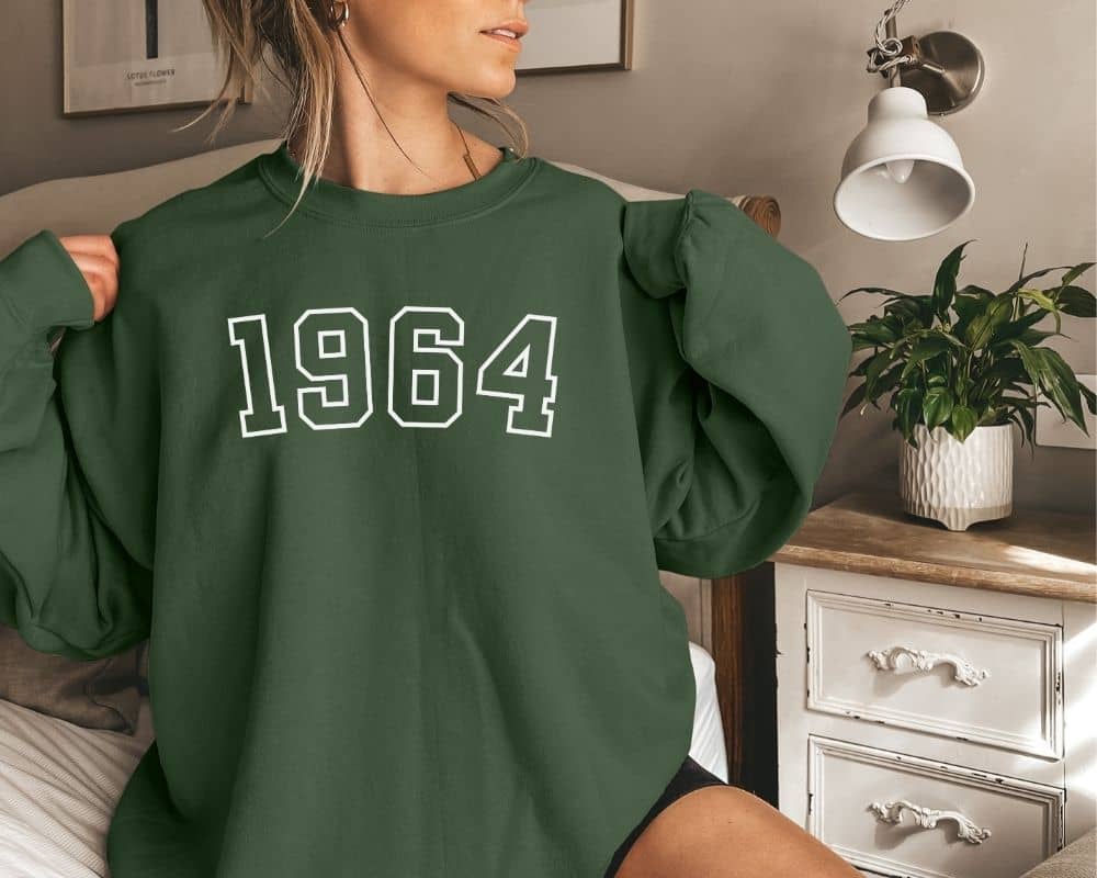 A person is wearing a forest green sweatshirt with '1964' printed on it, standing in a room with a dresser and a plant.