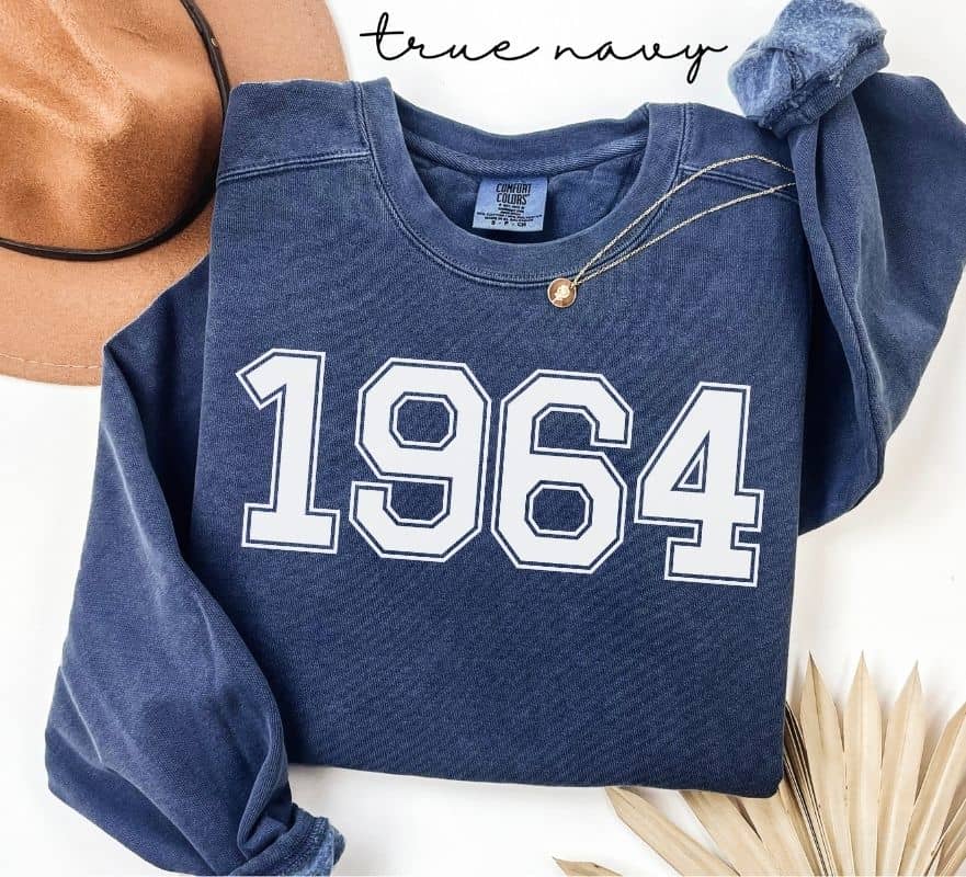 Blue (true navy) sweatshirt with '1964' printed on it, displayed with a hat and decorative elements.
