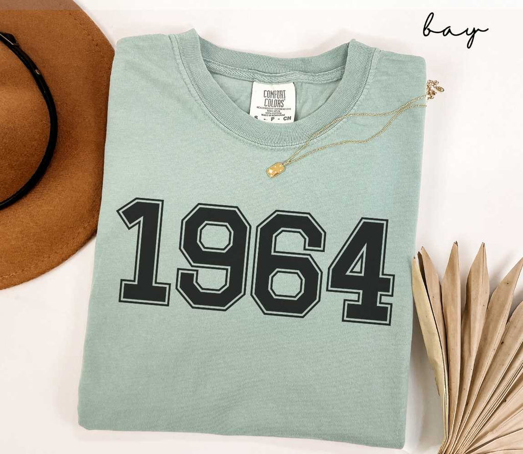 A light green (bay) t-shirt with "1964" printed on it, placed on a white surface alongside a hat and dried leaves.
