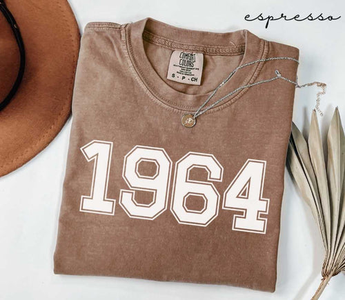 Folded brown (espresso) t-shirt with '1964' print on a white surface, accompanied by a hat and dried leaves.