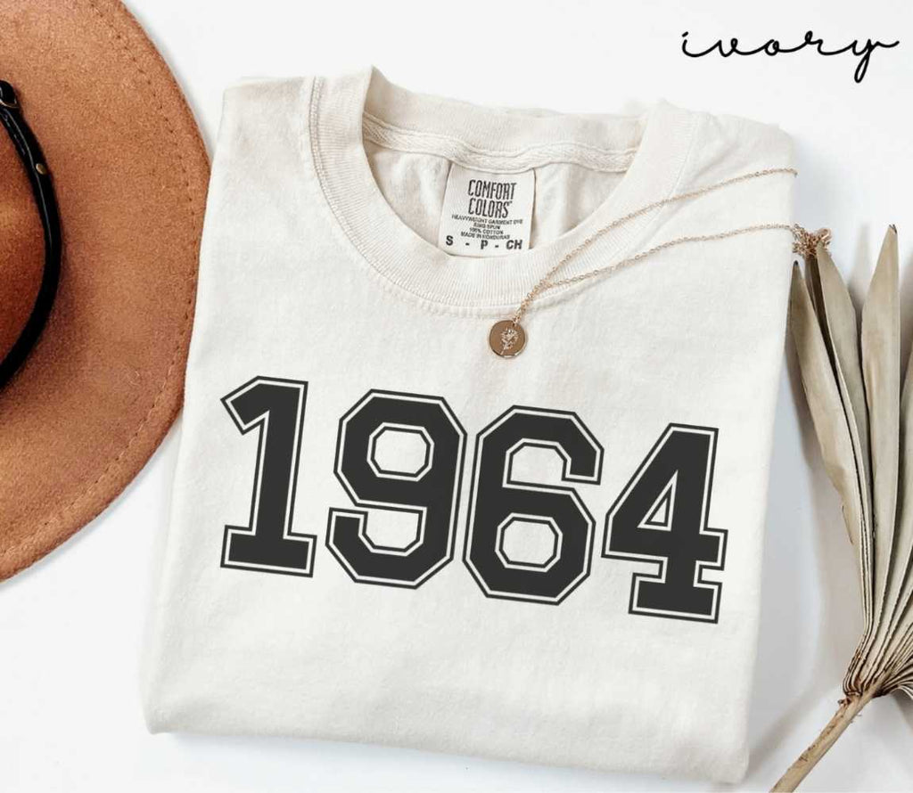 Beige (ivory) t-shirt with '1964' print, placed next to a brown hat and a dried plant on a light background.
