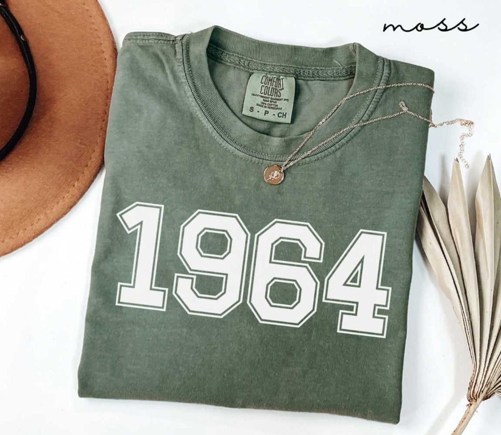Folded green (moss) shirt featuring a '1964' print on a white surface alongside a hat and dried leaves.
