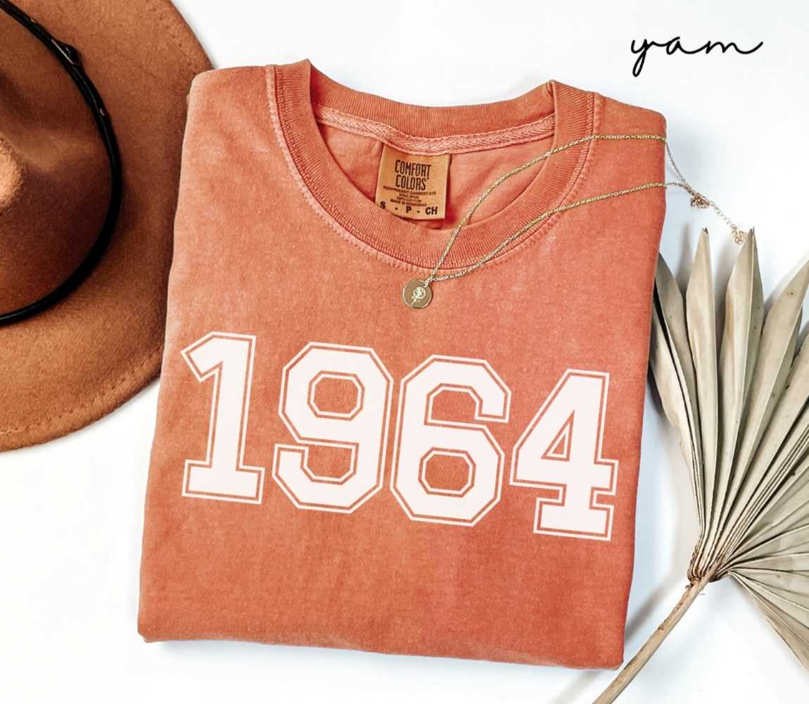 Folded orange (yam) t-shirt with a '1964' print, a brown hat, and a dried plant on a white background.