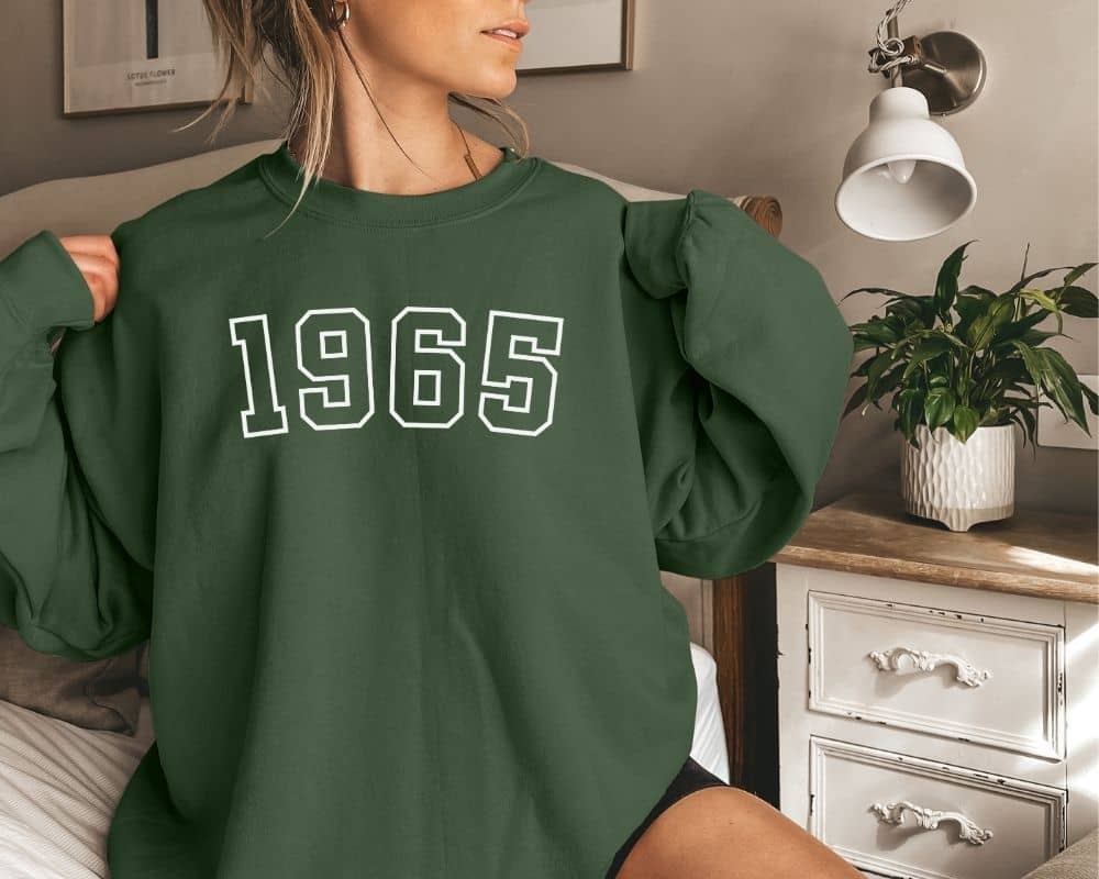 A person is wearing a green (forest green) sweatshirt that has '1965' printed on it while in a bedroom setting