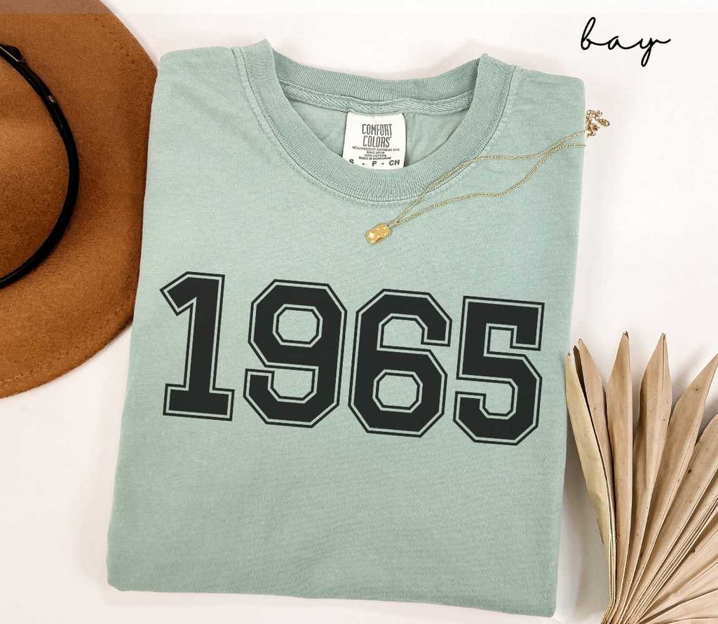 A light green t-shirt with '1965' printed on it, placed on a white surface, accompanied by a hat and dried leaves.
