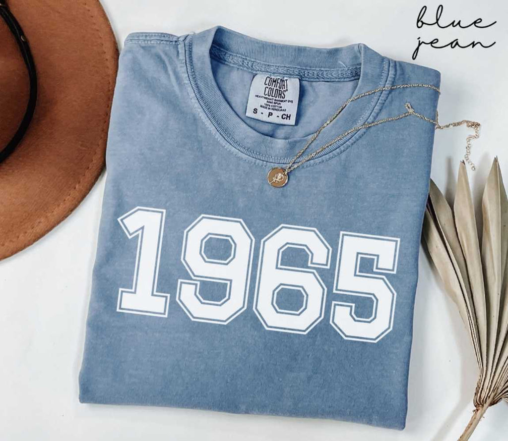 Folded blue jean t-shirt featuring '1963' print on a white surface, accompanied by a hat and dried leaves.