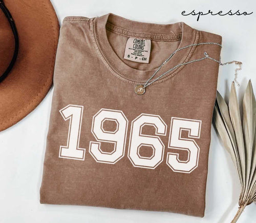 Folded brown (espresso) t-shirt with '1965' print on a white surface, accompanied by a hat and dried leaves.

