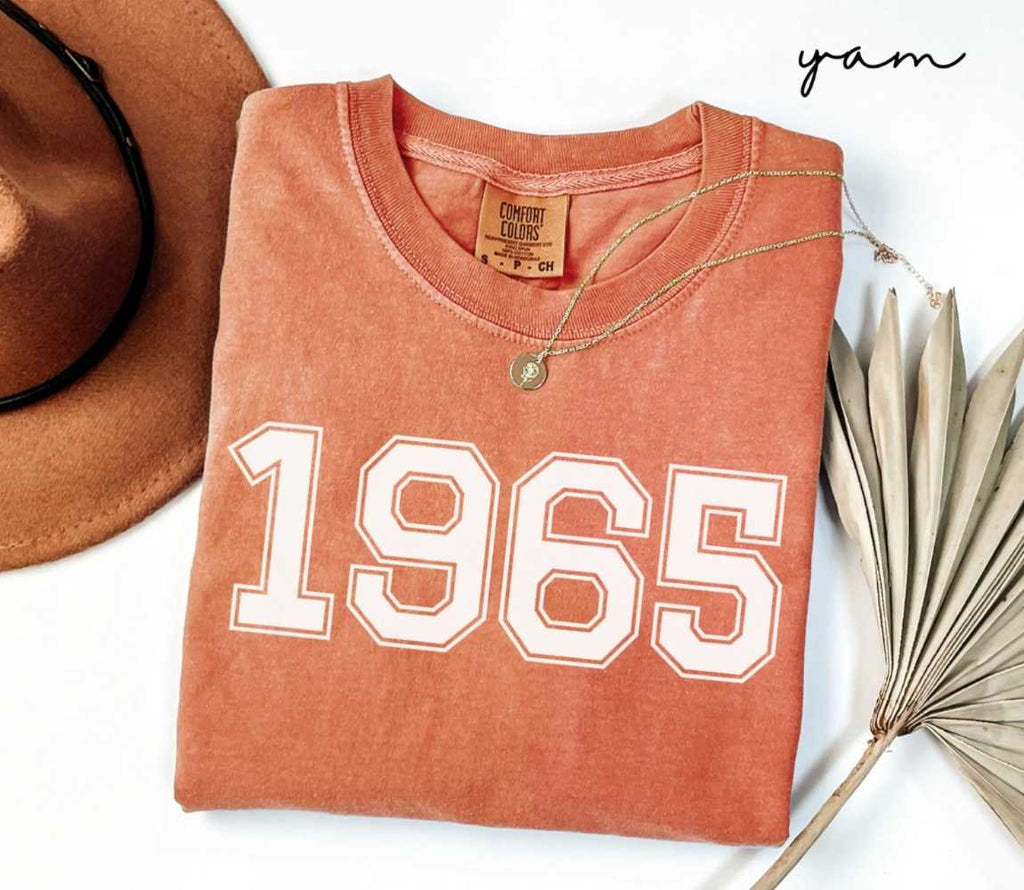 Folded orange (yam) t-shirt with a '1965' print, a brown hat, and a dried plant on a white background.
