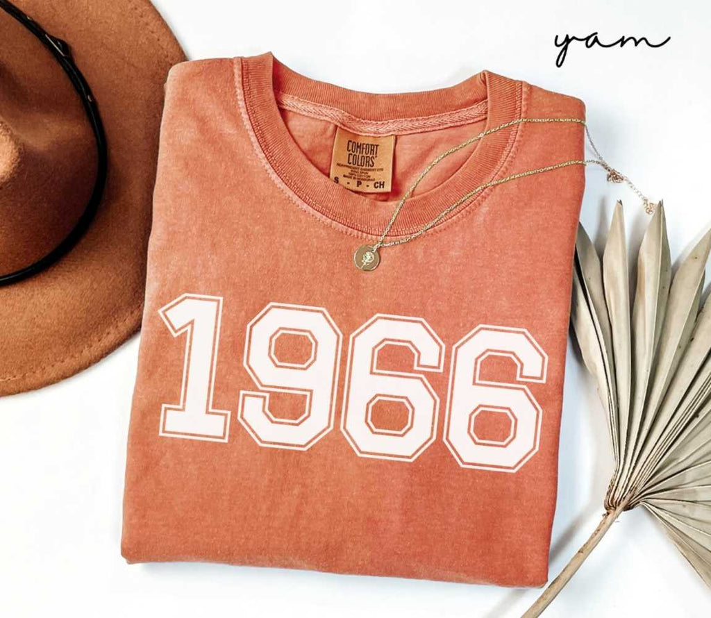 Folded orange (yam) t-shirt with a '1966' print, a brown hat, and a dried plant on a white background.
