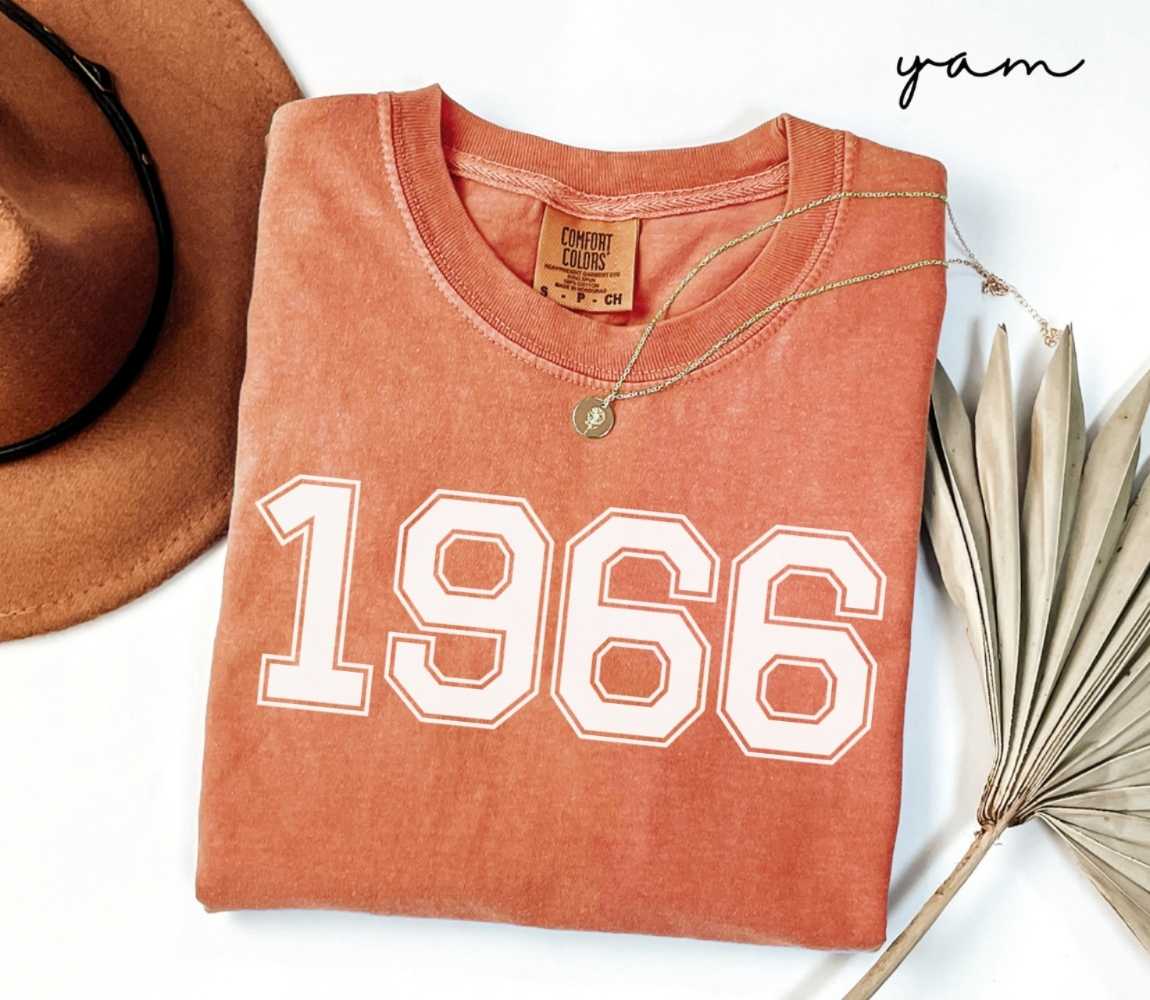 Folded orange (yam) t-shirt with a '1966' print, a brown hat, and a dried plant on a white background.

