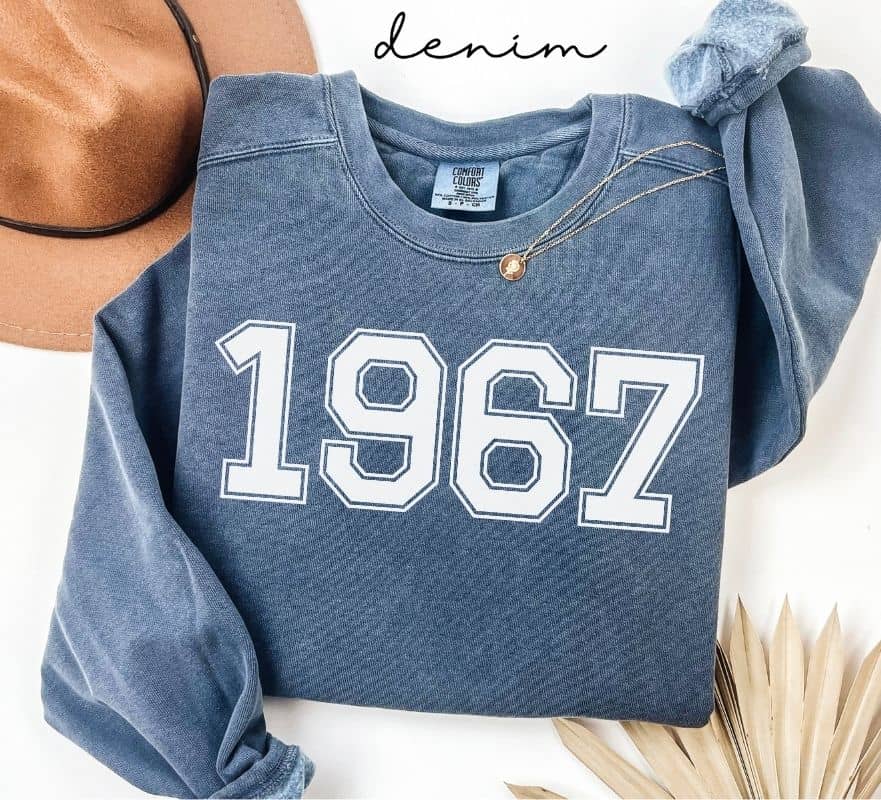 Blue (denim) sweatshirt with '1967' printed on it, worn by a person, with a plain background.