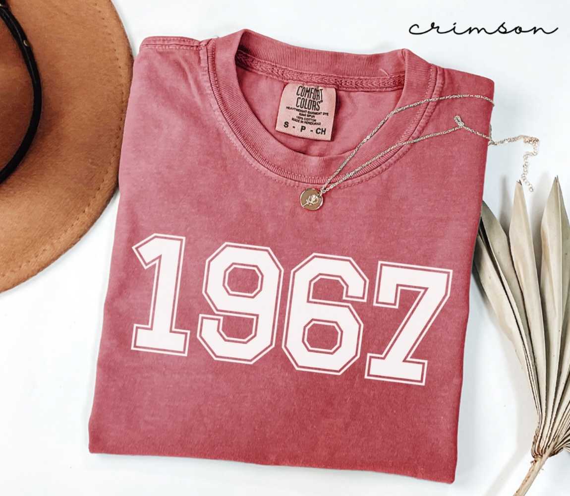A red (Crimson) folded t-shirt with '1967' printed on it, next to a hat, and dried leaves on a white background.