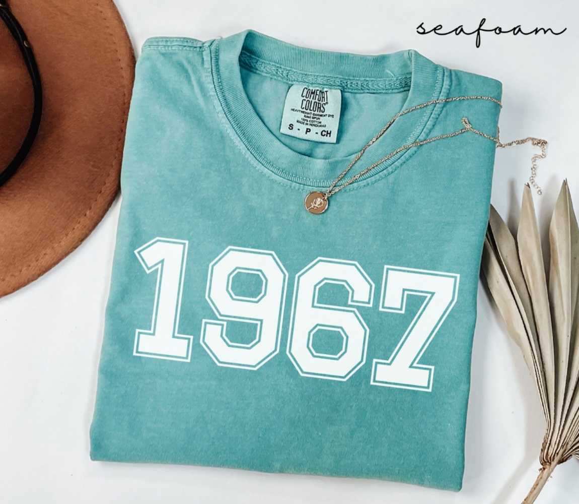 A folded (seafoam) teal t-shirt featuring a '1967' print lies next to a brown hat and dried leaves on a white background.