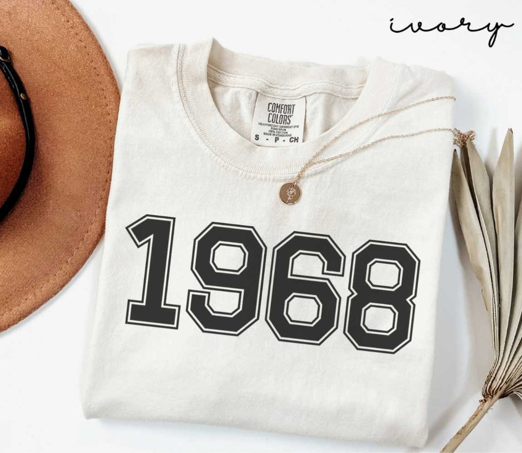 A beige (ivory) t-shirt with '1968' print, placed next to a brown hat and a dried plant on a light background.
