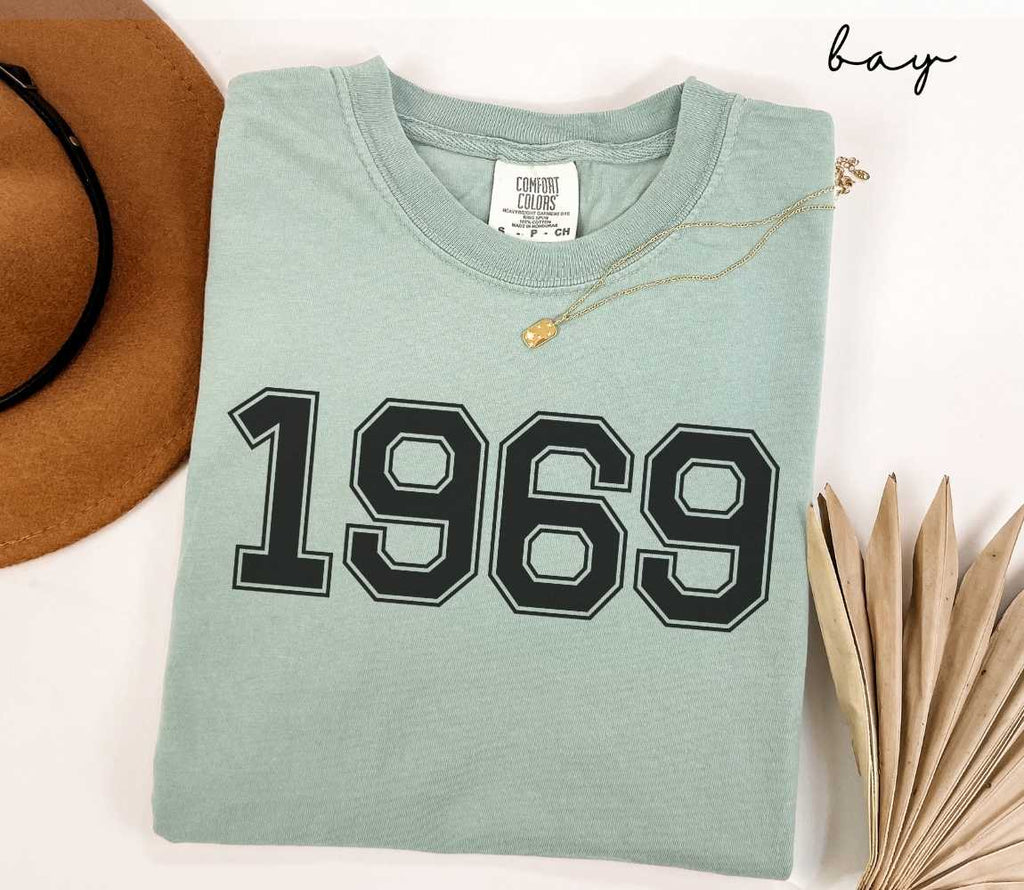 A light green (bay) t-shirt with "1969" printed on it, placed on a white surface alongside a hat and dried leaves.

