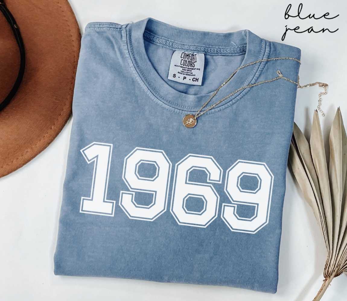 Folded blue jean t-shirt featuring '1969' print on a white surface, accompanied by a hat and dried leaves.
