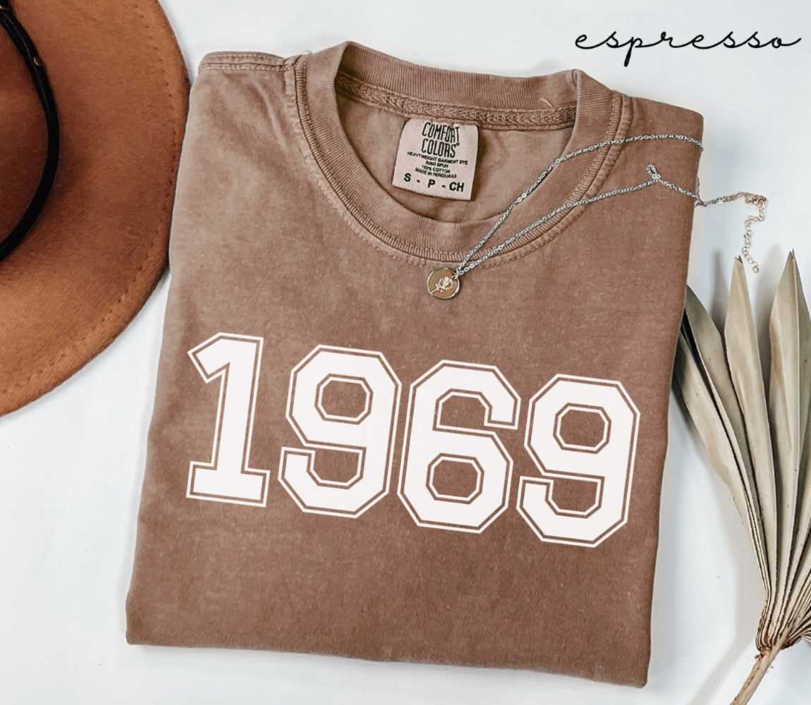 Folded brown (espresso) t-shirt with '1969' print on a white surface, accompanied by a hat and dried leaves.
