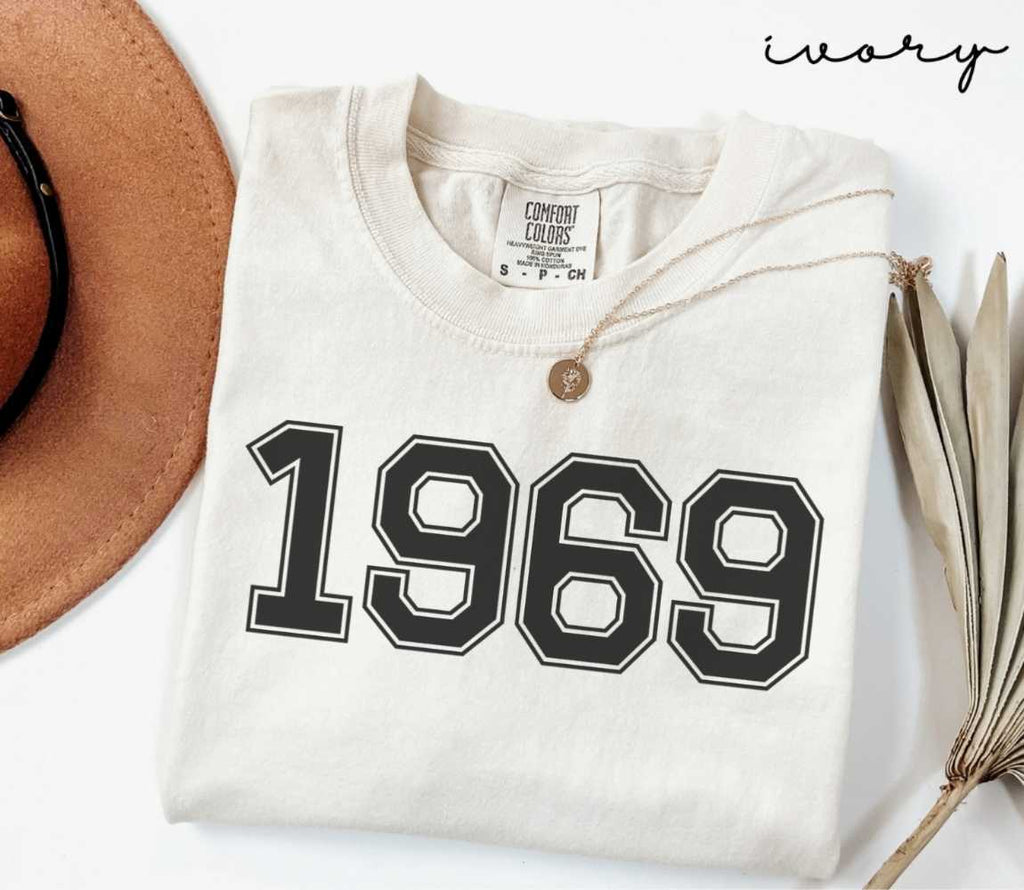 A beige (ivory) t-shirt with '1969' print, placed next to a brown hat and a dried plant on a light background.
