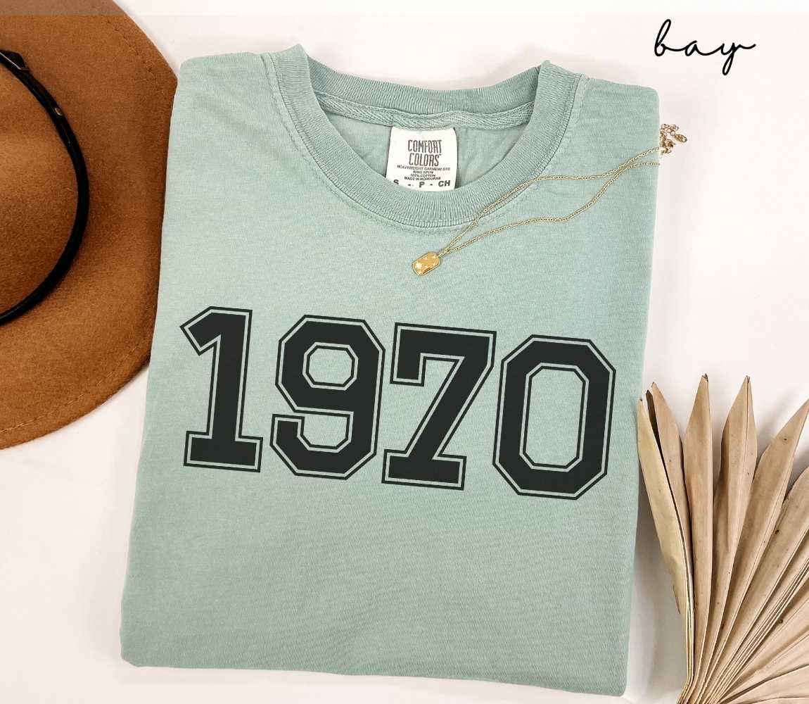 A light green (bay) t-shirt with "1970" printed on it, placed on a white surface alongside a hat and dried leaves.
