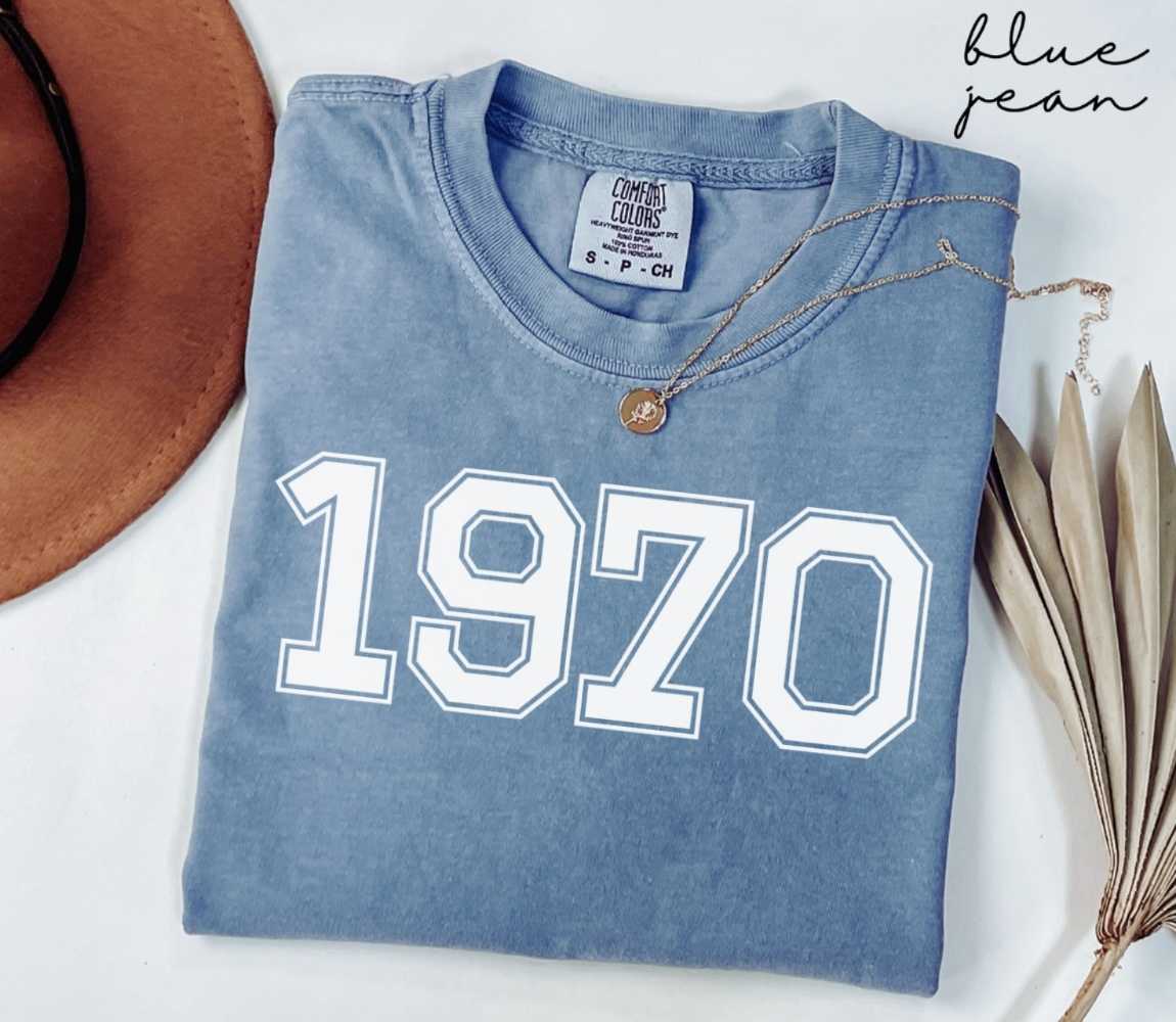 Folded blue jean t-shirt featuring '1970' print on a white surface, accompanied by a hat and dried leaves.

