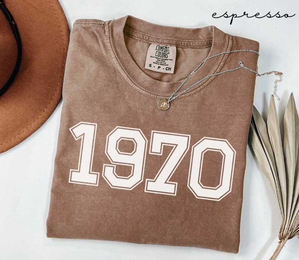 Folded brown (espresso) t-shirt with '1970' print on a white surface, accompanied by a hat and dried leaves.
