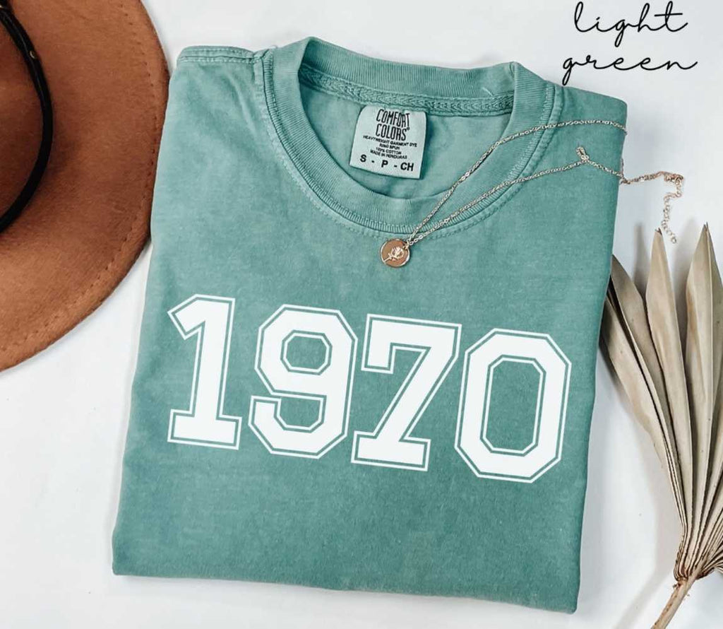 A light green t-shirt with '1970' printed on it, placed on a white surface, accompanied by a hat and dried leaves.

