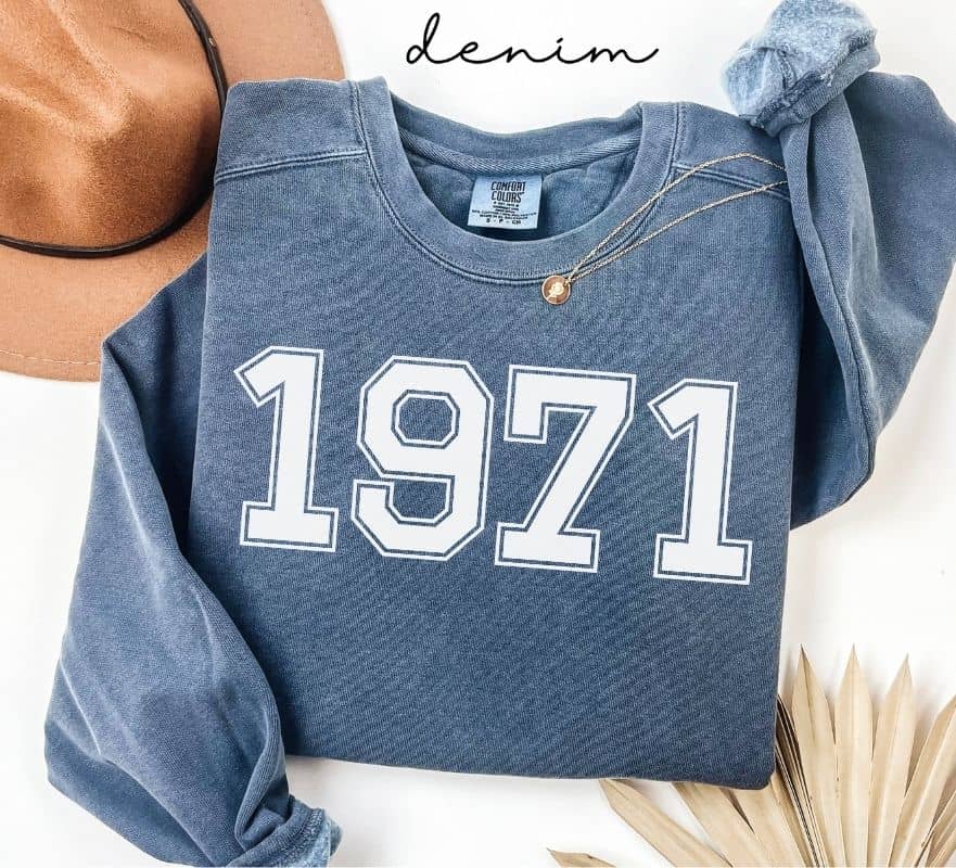 Blue (denim) sweatshirt with '1971' printed on it, worn by a person, with a plain background.