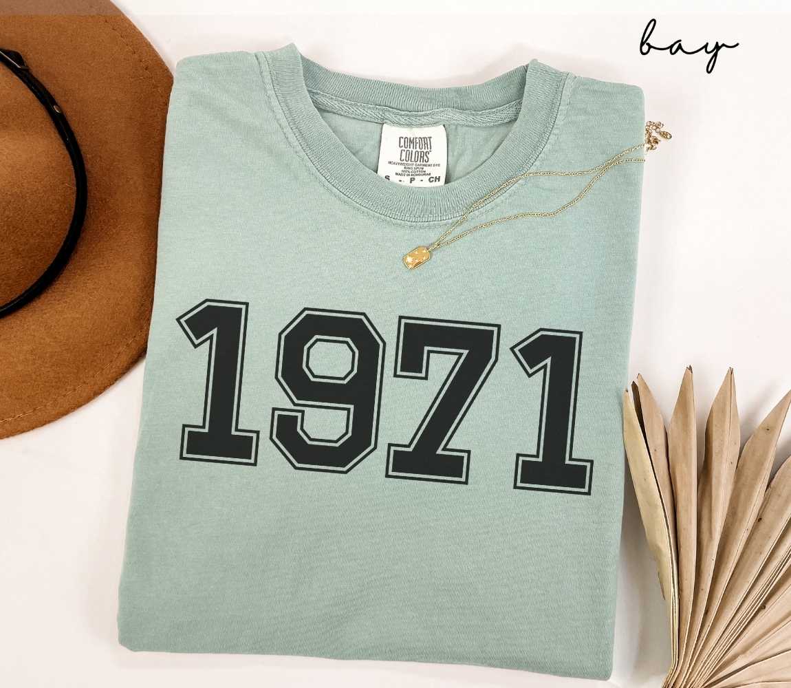 A light green (bay) t-shirt with "1971" printed on it, placed on a white surface alongside a hat and dried leaves.
