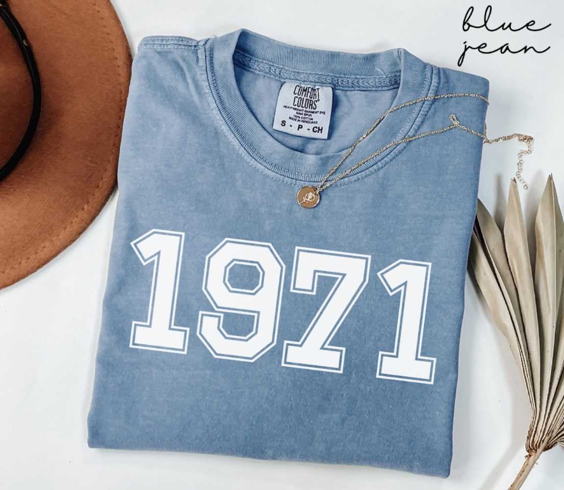 Folded blue jean t-shirt featuring '1971' print on a white surface, accompanied by a hat and dried leaves.
