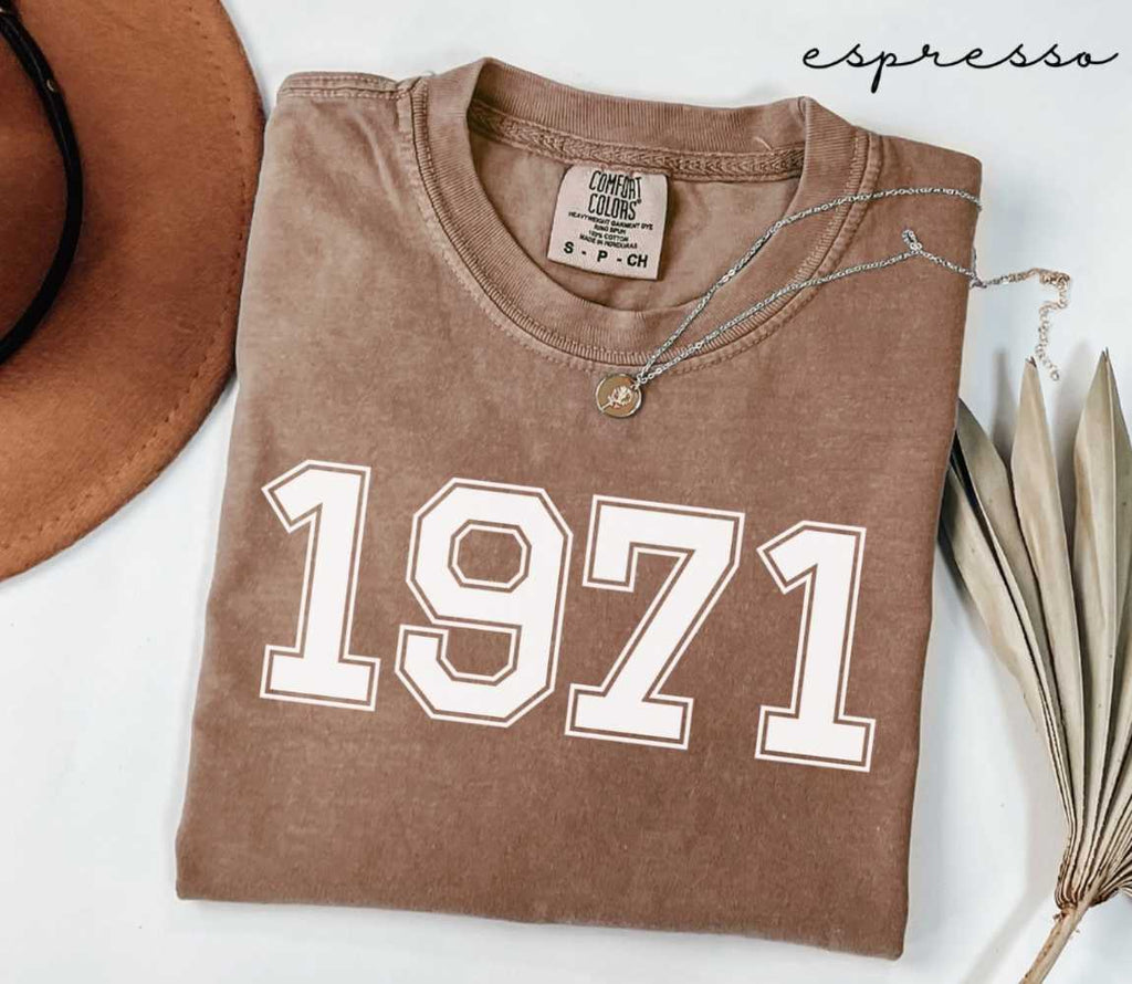 Folded brown (espresso) t-shirt with '1971' print on a white surface, accompanied by a hat and dried leaves.
