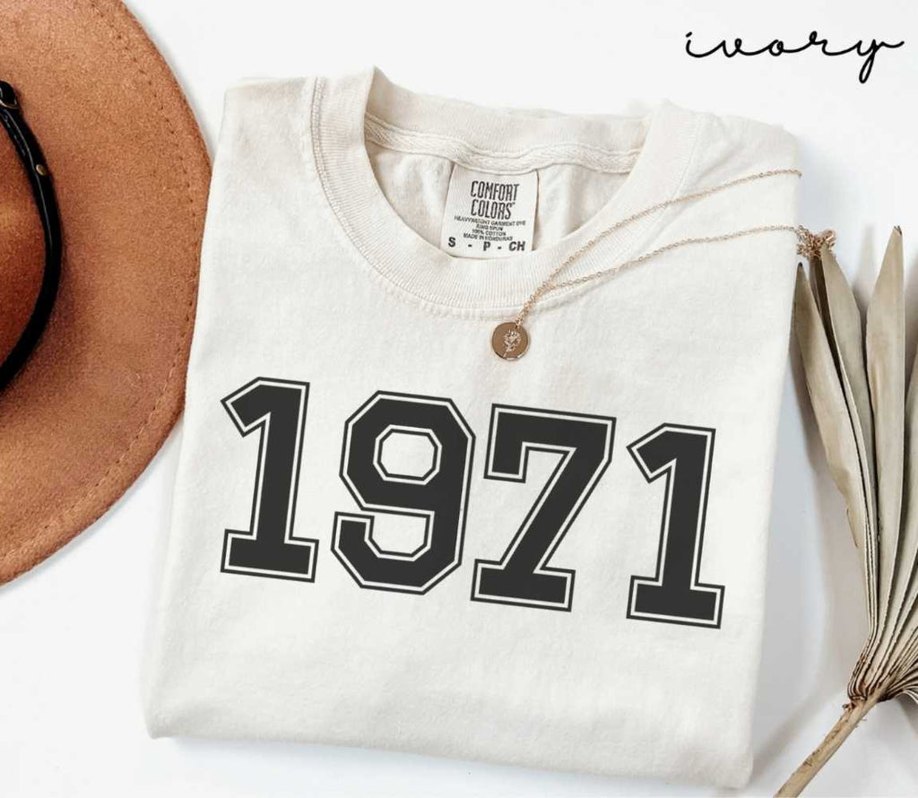 A beige (ivory) t-shirt with '1971' print, placed next to a brown hat and a dried plant on a light background.
