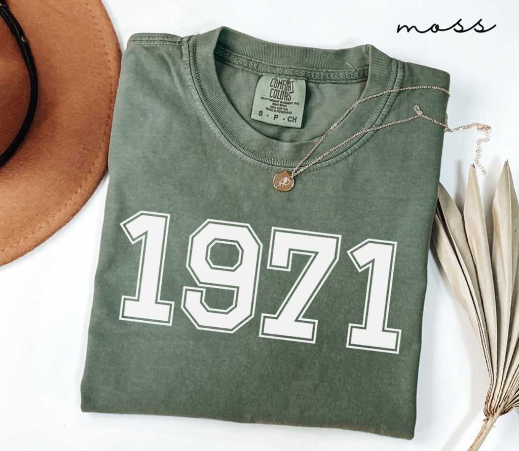 Folded green (moss) t-shirt featuring a '1971' print on a white surface alongside a hat and dried leaves.
