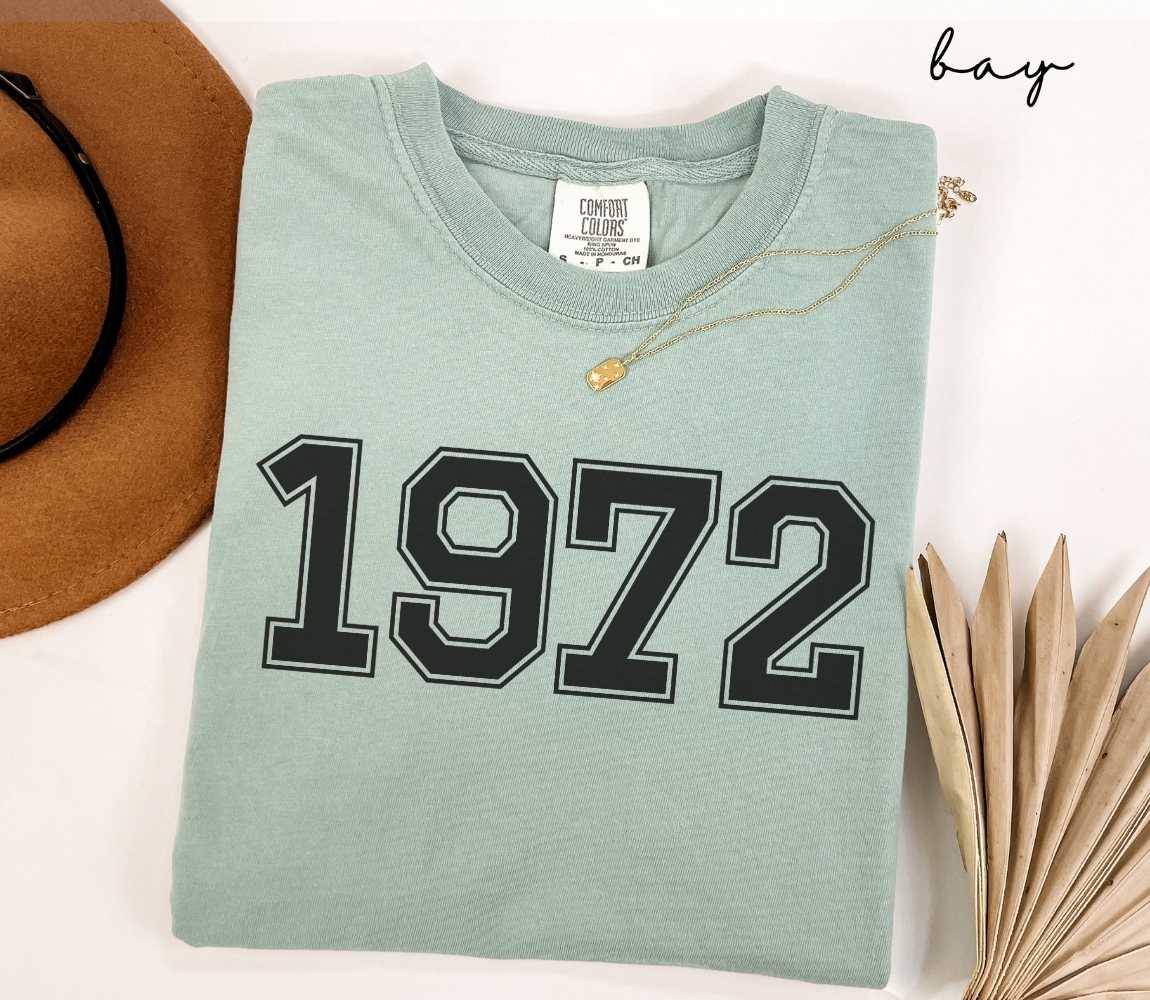 A light green (bay) t-shirt with "1972" printed on it, placed on a white surface alongside a hat and dried leaves.

