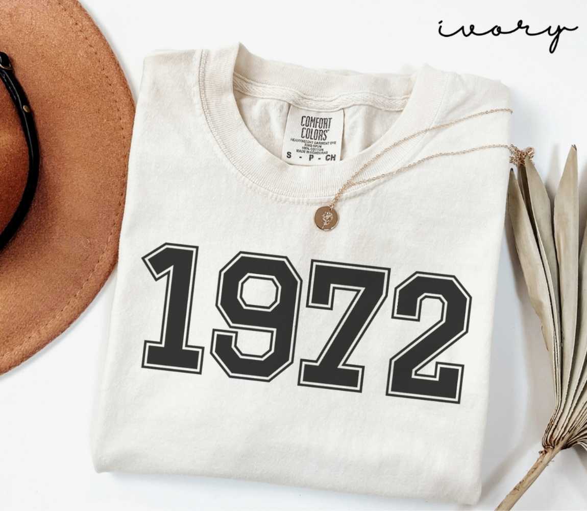 A beige (ivory) t-shirt with '1972' print, placed next to a brown hat and a dried plant on a light background.
