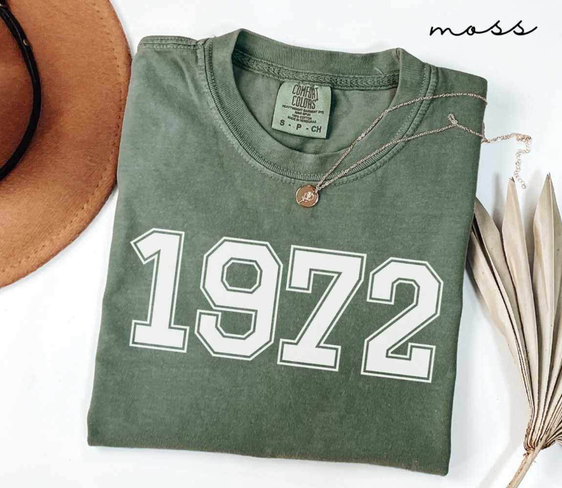 Folded green (moss) t-shirt featuring a '1972' print on a white surface alongside a hat and dried leaves.
