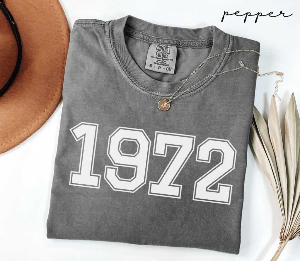 Folded gray (pepper) t-shirt with '1972' print on a white surface with a hat and dried leaves.
