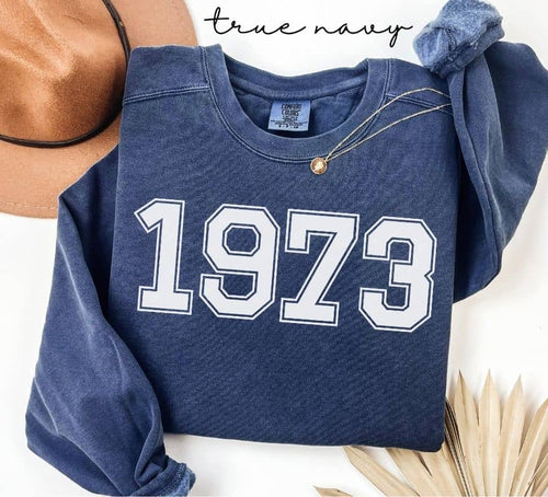 Blue (true navy) sweatshirt with '1973' printed on it, displayed with a hat and decorative elements.