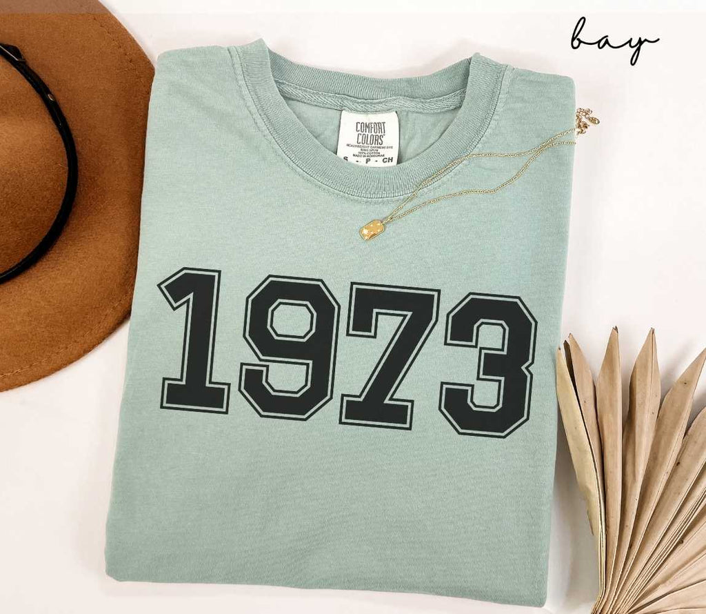 A light green t-shirt with '1973' printed on it, placed on a white surface, accompanied by a hat and dried leaves.
