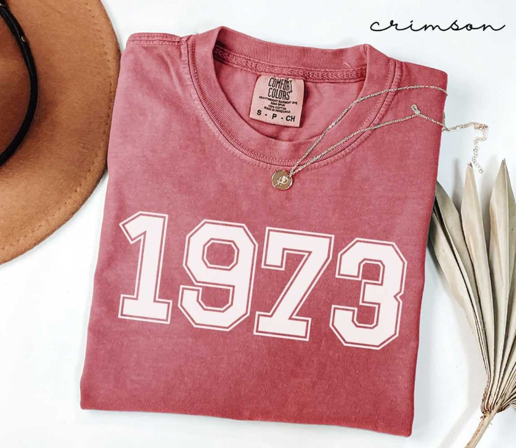 A red (Crimson) folded t-shirt with '1973' printed on it, next to a hat, and dried leaves on a white background.
