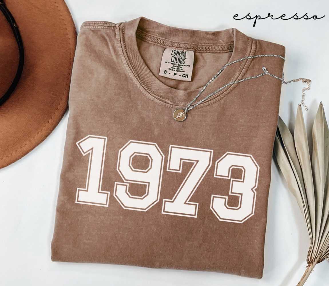 Folded brown (espresso) t-shirt with '1973' print on a white surface, accompanied by a hat and dried leaves.
