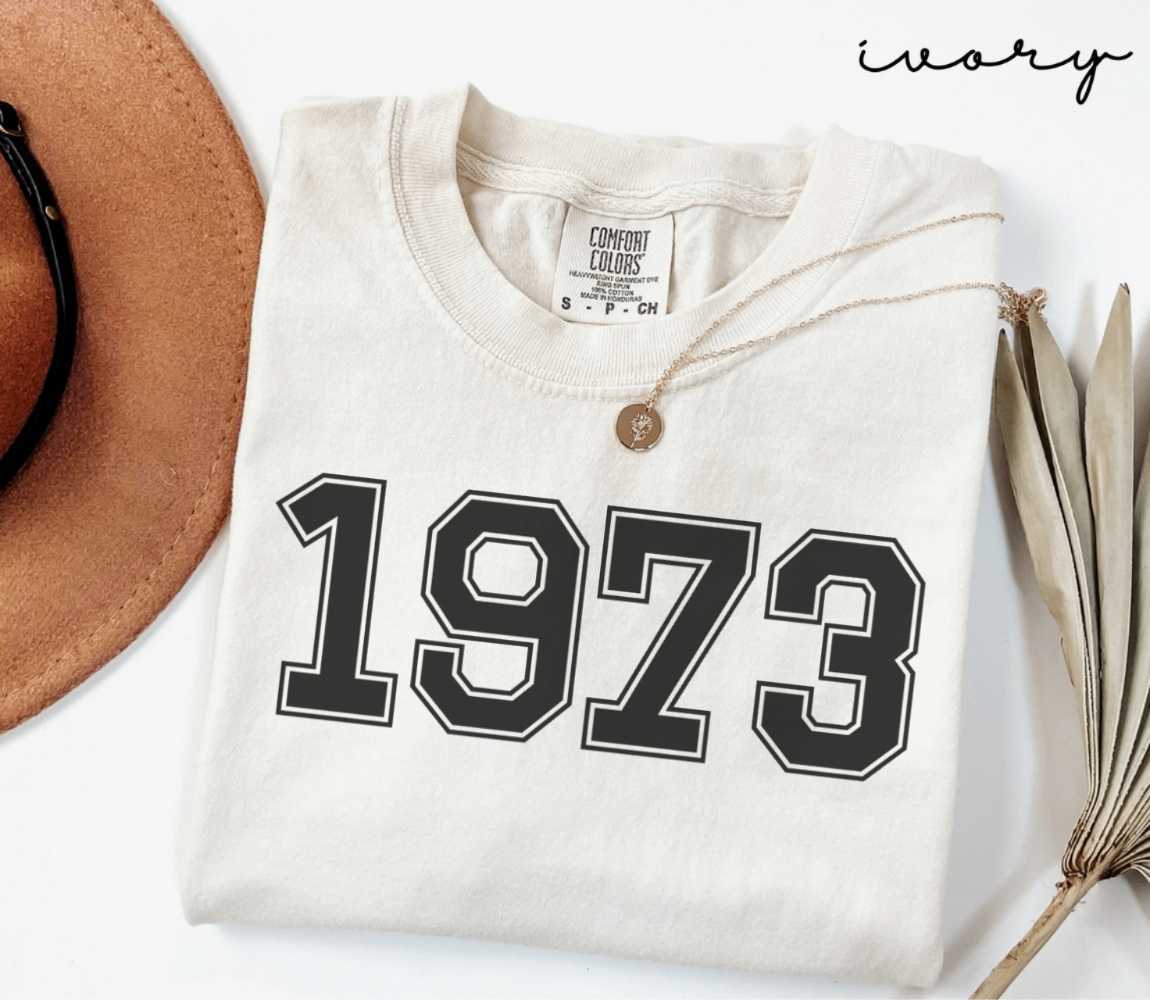 A beige (ivory) t-shirt with '1973' print, placed next to a brown hat and a dried plant on a light background.