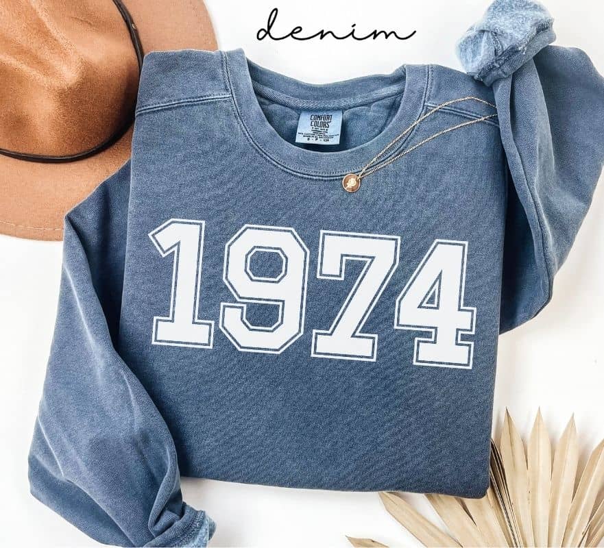 Blue (denim) sweatshirt with '1974' printed on it, worn by a person, with a plain background.