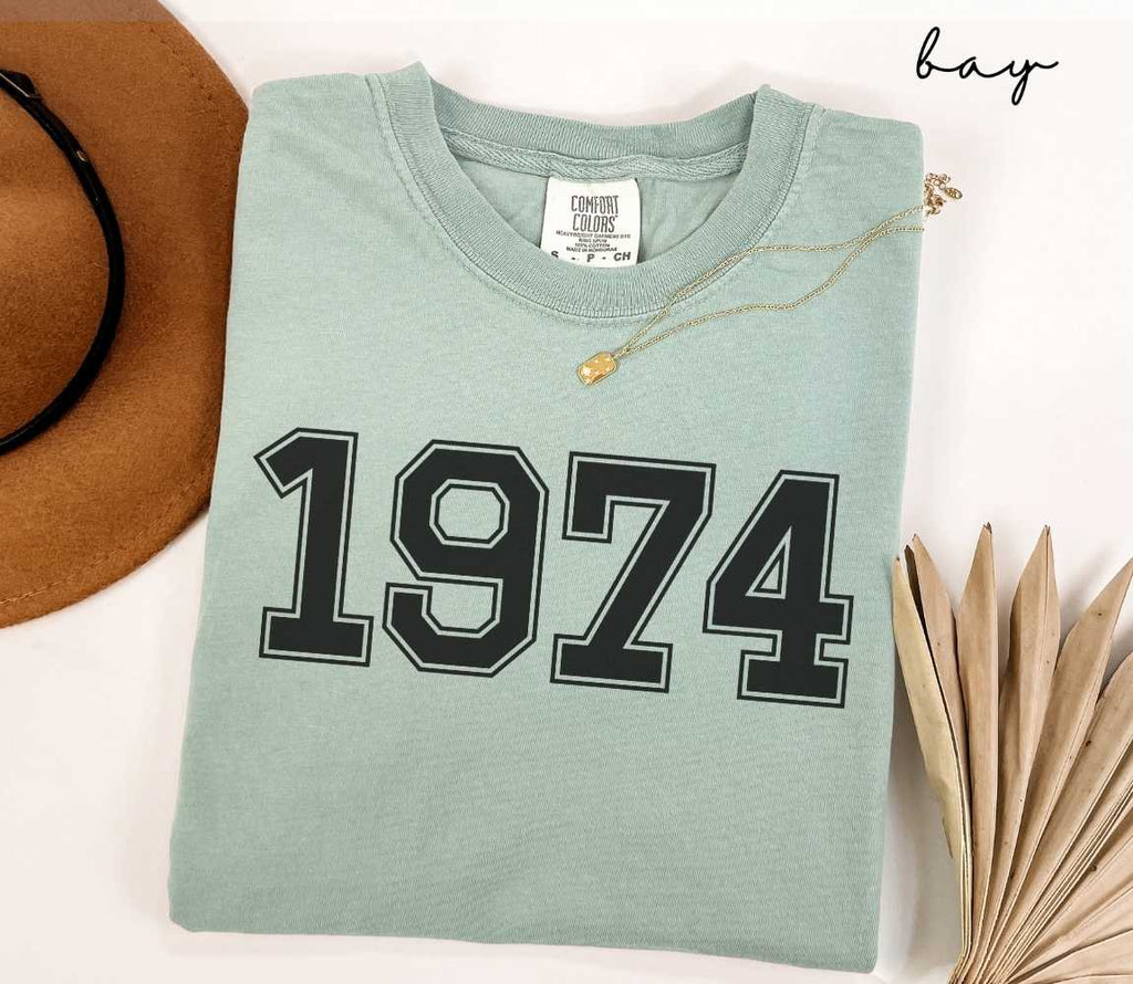 A light green (bay) t-shirt with "1974" printed on it, placed on a white surface alongside a hat and dried leaves.