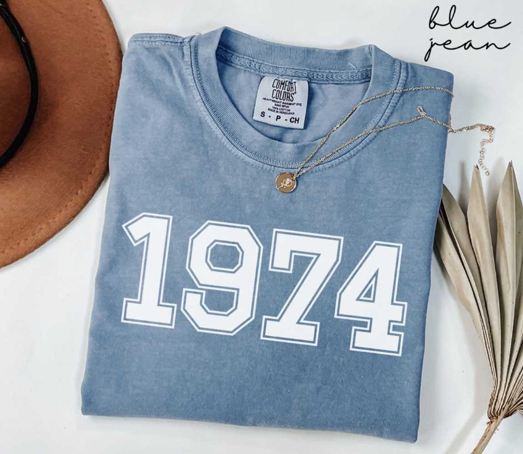 Folded blue jean t-shirt featuring '1974' print on a white surface, accompanied by a hat and dried leaves.
