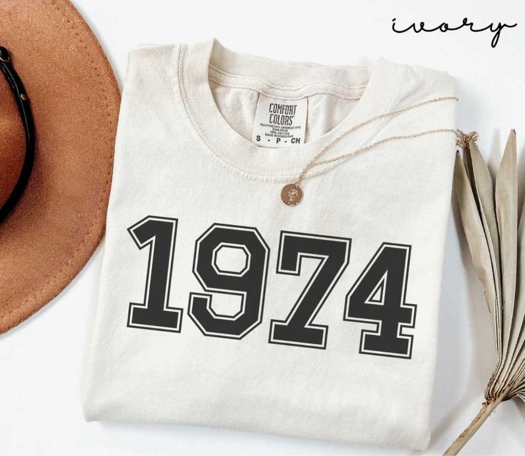A beige (ivory) t-shirt with '1974' print, placed next to a brown hat and a dried plant on a light background.
