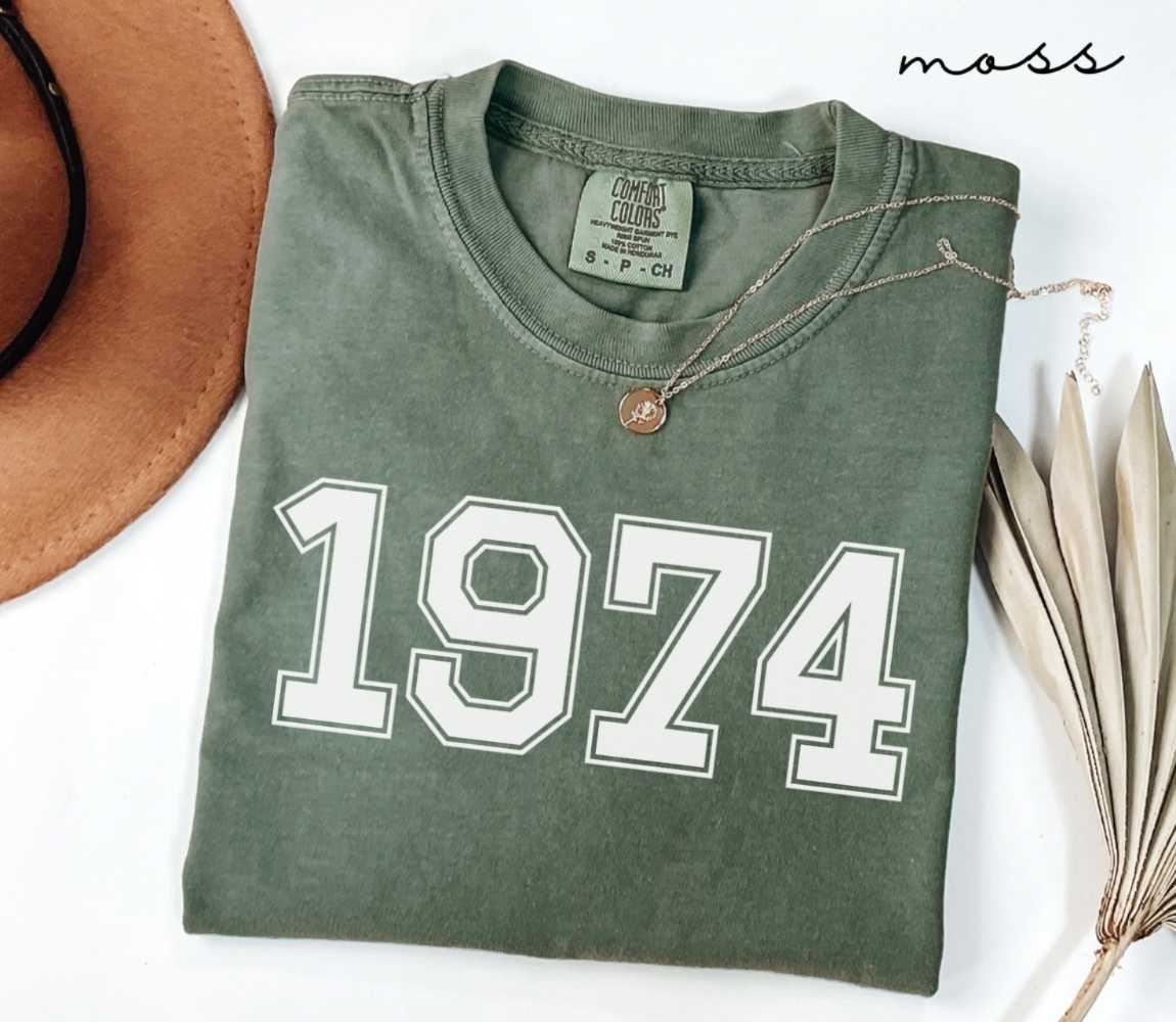Folded green (moss) t-shirt featuring a '1974' print on a white surface alongside a hat and dried leaves.
