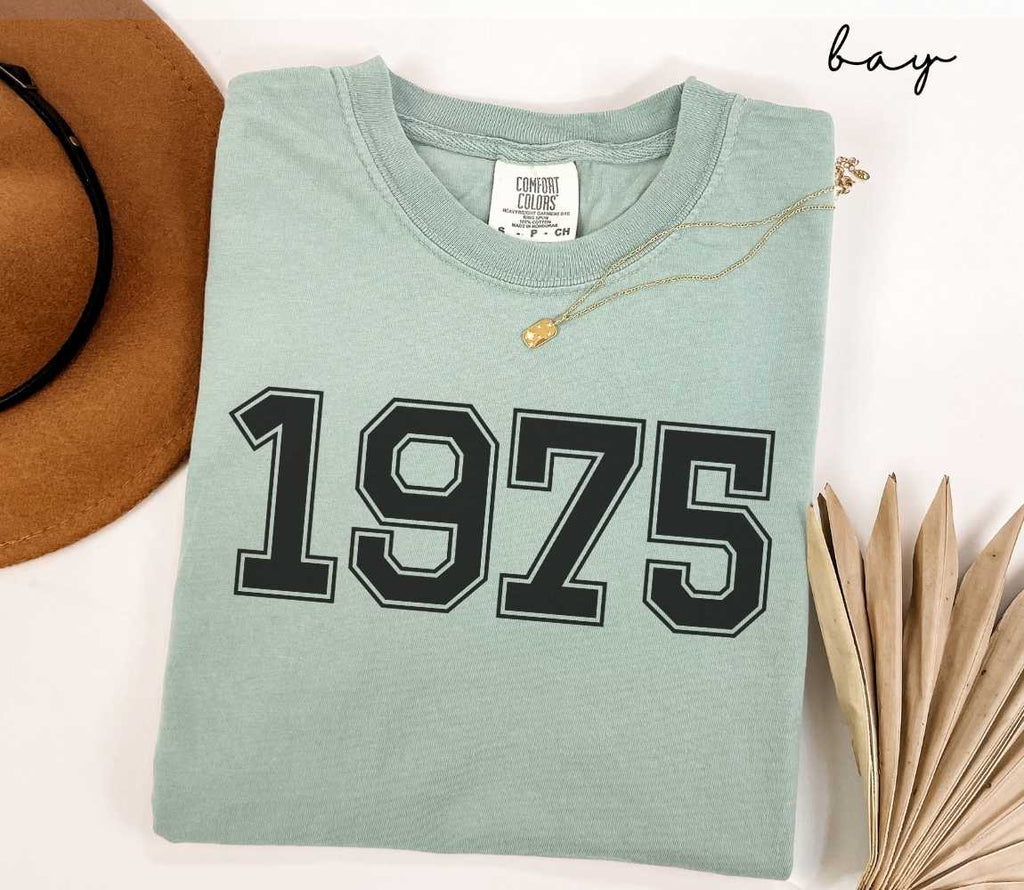 A light green (bay) t-shirt with "1975" printed on it, placed on a white surface alongside a hat and dried leaves.
