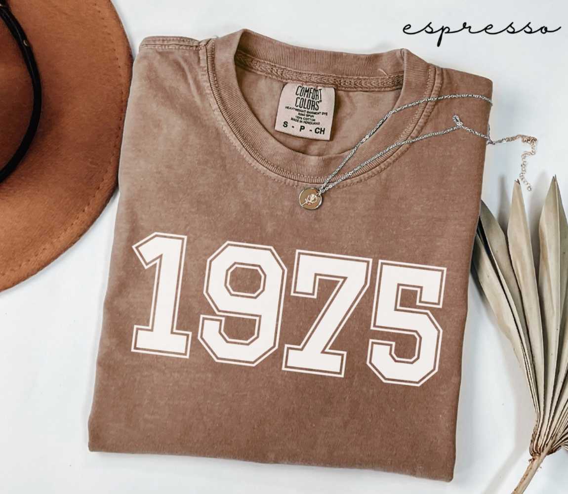 Folded brown (espresso) t-shirt with '1975' print on a white surface, accompanied by a hat and dried leaves.
